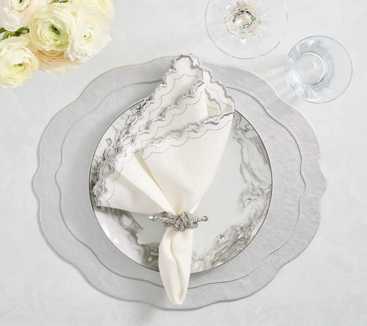 A chic table setting features the Kim Seybert Tailored Placemat Set of 4 in Silver, paired with scalloped plates, marble-patterned inner plate, jeweled napkin holder, and clear wine and water glasses beside a bouquet of white flowers.