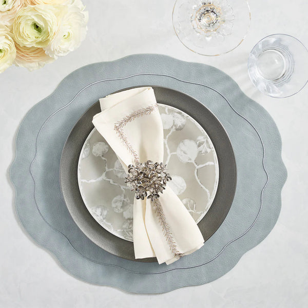 Elegant table setting featuring the Kim Seybert Tailored Placemat Set of 4 in Steel Grey, paired with a silver charger, patterned plate, cream napkin with ring, crystal wine and water glasses, and white ranunculus flowers in the upper left.
