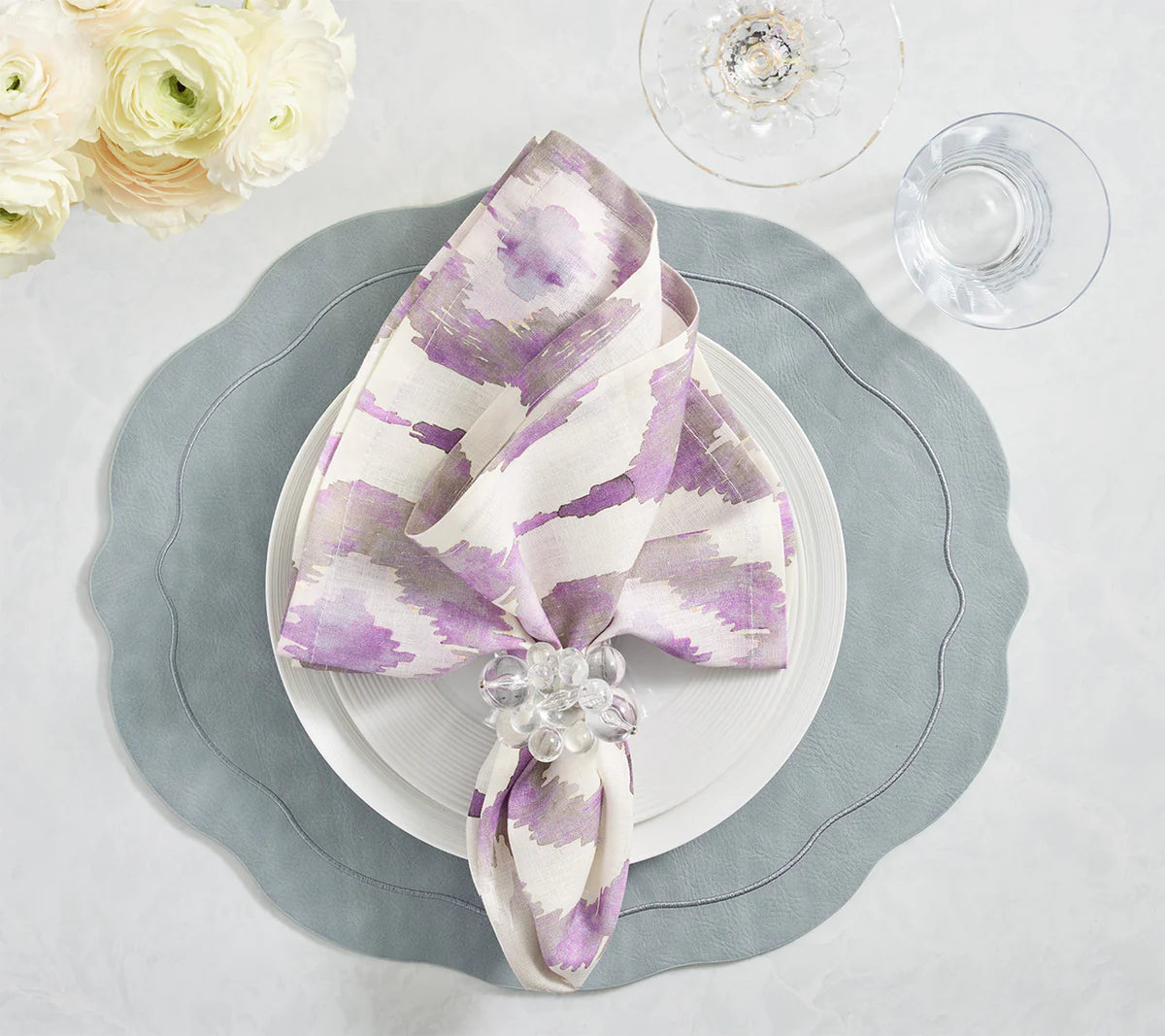 A white plate sits on the Kim Seybert Tailored Placemat in Steel Grey, topped with a white and purple napkin in a glass ring. Nearby are a wine glass, water glass, and white flowers.