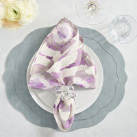 An elegant table is set with a scalloped gray placemat, white plates, and a lavender-patterned napkin held by the Kim Seybert Cloud Napkin Ring Set of 4. Glassware, acrylic baubles, and white flowers complete the look.