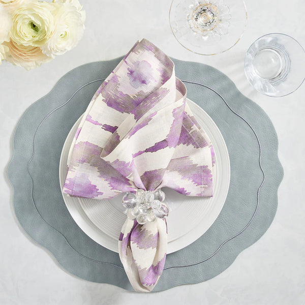 An elegant table is set with a scalloped gray placemat, white plates, and a lavender-patterned napkin held by the Kim Seybert Cloud Napkin Ring Set of 4. Glassware, acrylic baubles, and white flowers complete the look.