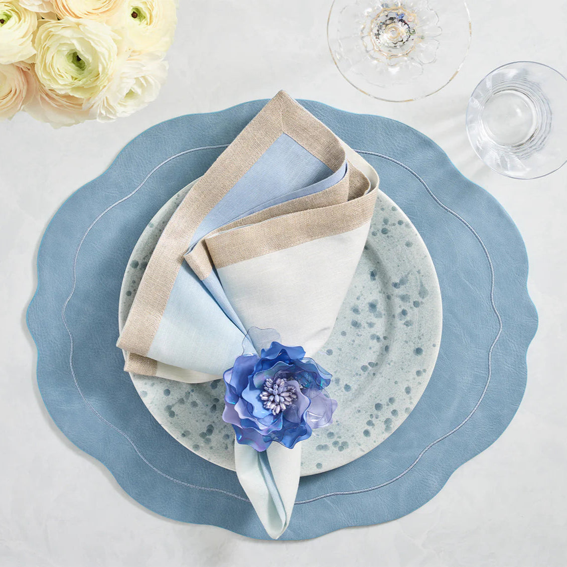 Elegant table setting featuring Kim Seybert Dip Dye Napkins - Cool Tones (Set of 4), a blue scalloped placemat, speckled ceramic plate, purple flower napkin ring, glassware, and white flowers in the background.