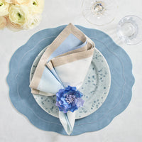 Elegant table setting featuring Kim Seybert Dip Dye Napkins - Cool Tones (Set of 4), a blue scalloped placemat, speckled ceramic plate, purple flower napkin ring, glassware, and white flowers in the background.