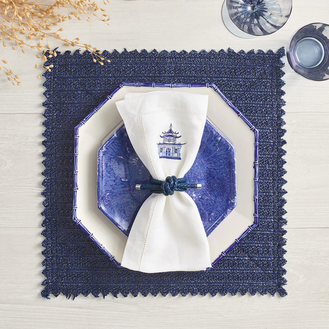 A blue octagonal plate rests on a cream square plate atop a navy textured placemat. A white napkin with a blue pagoda print is held by the Kim Seybert Rope Knot Napkin Ring Set of 4, with dried flowers and glassware nearby on a light wood table.