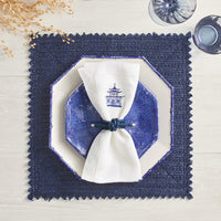 A blue octagonal plate rests on a cream square plate atop a navy textured placemat. A white napkin with a blue pagoda print is held by the Kim Seybert Rope Knot Napkin Ring Set of 4, with dried flowers and glassware nearby on a light wood table.