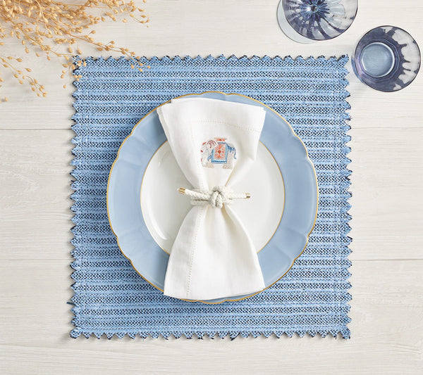 A table is set with a Kim Seybert Vista Reversible Placemat in Navy & Periwinkle, a white plate with blue rim, a knotted embroidered napkin, two blue glasses on light wood, and dried plants in the top left corner.