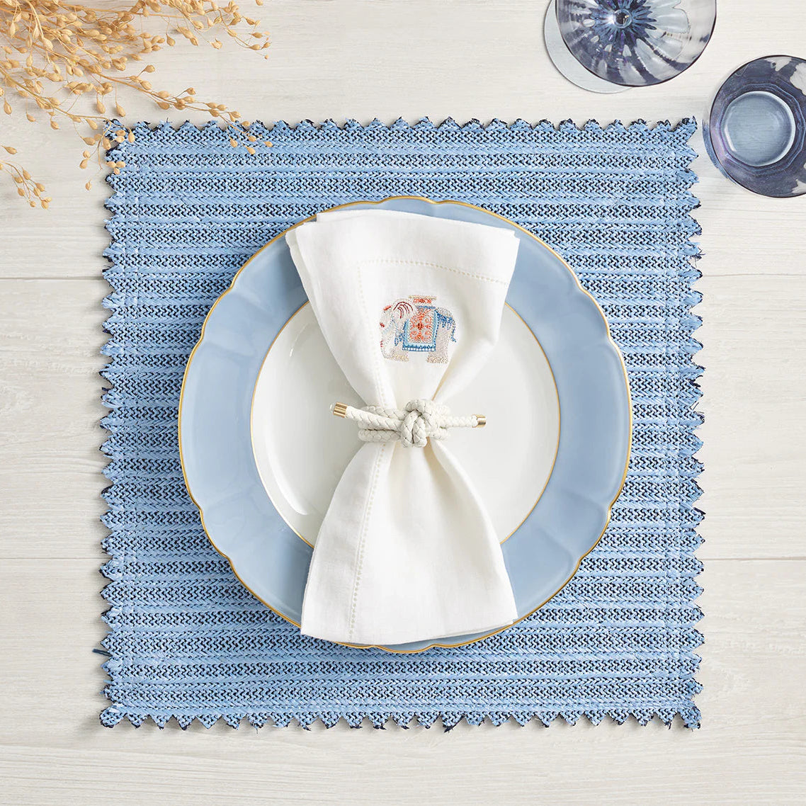 A blue faux leather placemat with a white plate featuring a light blue border is topped with a white cloth napkin and the Rope Knot Napkin Ring Set of 4 by Kim Seybert. Blue water glasses and dried twigs accent this coastal table on light wood.