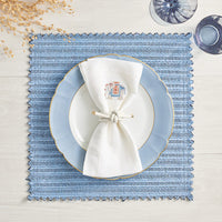 A blue faux leather placemat with a white plate featuring a light blue border is topped with a white cloth napkin and the Rope Knot Napkin Ring Set of 4 by Kim Seybert. Blue water glasses and dried twigs accent this coastal table on light wood.