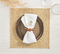 A brown plate rests on a Kim Seybert Vista Reversible Placemat in ivory & beige, with a white napkin featuring an embroidered pineapple and brown napkin ring. Two clear glasses and dried foliage sit on the light wooden table.