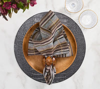 A wooden bowl holding a Windsor Napkin from the Kim Seybert Napkin Set of 4 - Multi rests on a black textured charger plate, accompanied by two clear glasses with gold rims atop a white marble surface; neutral tones and hints of purple flowers can be seen in the corner.