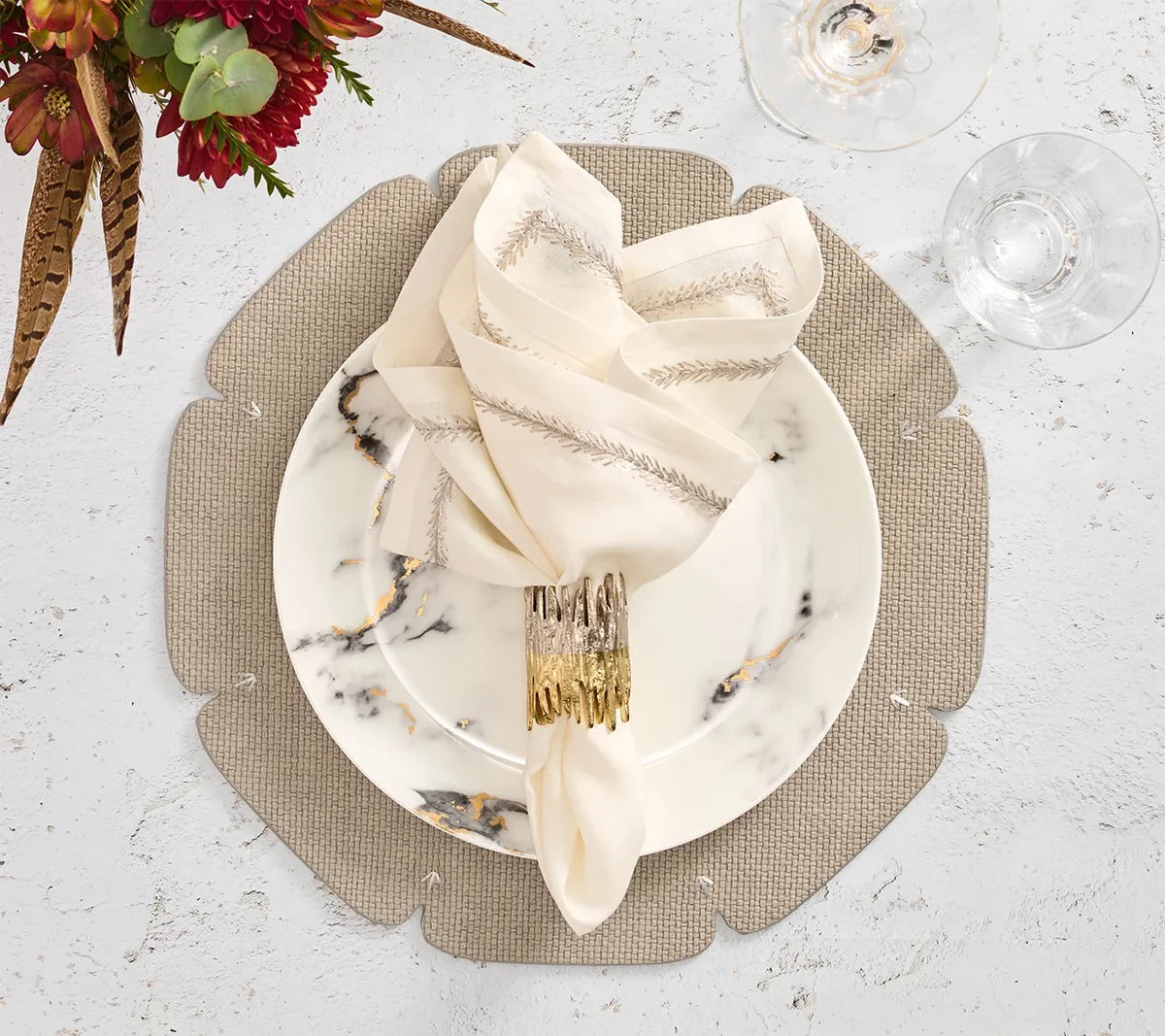 A marble-patterned plate with a gold-rimmed napkin holder and neatly folded cream napkin rests on the Kim Seybert Divot Placemat Set of 4 in Grey; nearby, wine glasses and a floral arrangement accent the white tabletop.