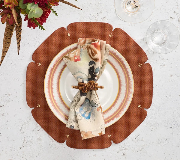 A floral napkin with a woven ring rests on stacked plates atop the Kim Seybert Divot Placemat Set of 4 in brown, a chic and modern dining accessory, flanked by two empty wine glasses on a white textured surface.