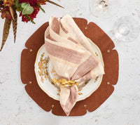 A gold and white plate with a folded striped napkin and gold napkin ring rests on the Kim Seybert Divot Placemat Set of 4 in Brown, adding an elegant touch to the dining table. Two glasses and a flower placed nearby on a white surface complete the modern yet inviting table setting.