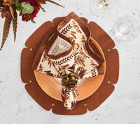 A fall-themed table setting featuring a patterned napkin in a gold ring on an orange plate is complemented by the contemporary Kim Seybert Divot Placemat Set of 4 in Brown, set next to wine glasses and autumn flowers.