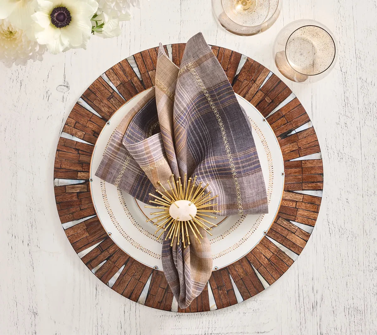 Top view of a Current Home NY Halo Placemat set with a brown plaid napkin, gold burst napkin ring, clear glasses, and a flower on a white wooden table.