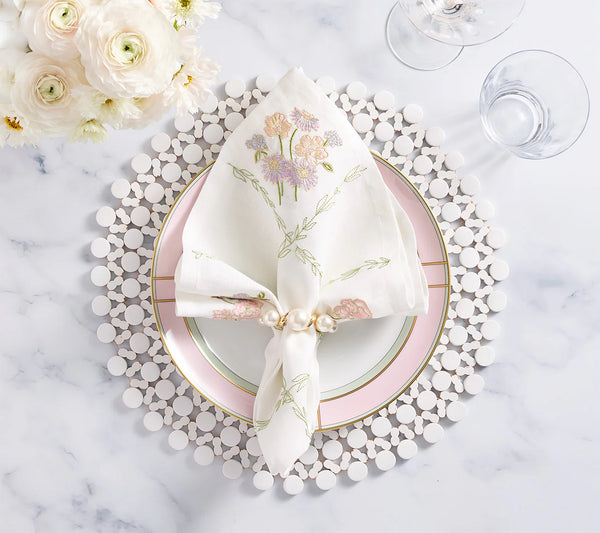 Elegant table setting with a white plate edged in pink and gold, topped with a floral embroidered napkin held by the Kim Seybert Pearl Drift Napkin Ring Set of 4 in Pearl & Gold. Clear glasses and white flowers rest on a marble surface.