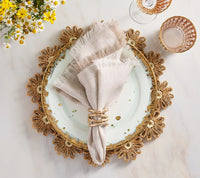 A white plate with gold accents sits on a floral gold placemat, creating charming farmhouse table decor. Resting on the plate is a Kim Seybert Chambray Fringe Napkin from the Set of 4, neatly placed in a woven ring. Nearby, crystal glasses and a yellow and white flower bouquet enhance the elegance of the marble table setting.