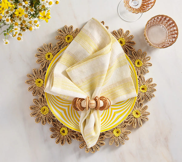A coastal table features a yellow and white striped plate on a daisy placemat, topped with a Kim Seybert Broad Stripe Napkin (set of 4) in a wooden ring. Pink crystal glasses and daisy blooms complete the look.