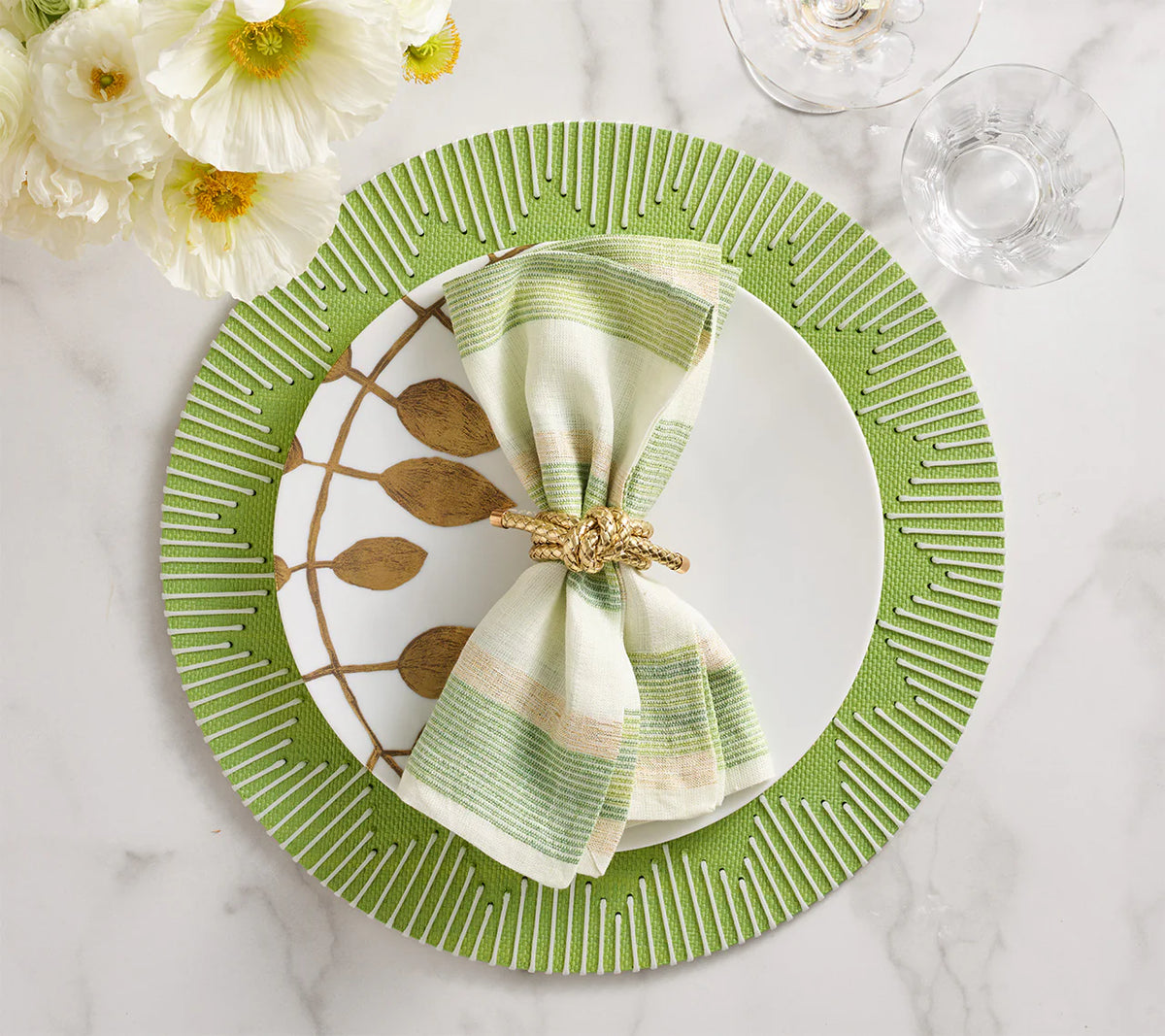 A white plate sits on a Kim Seybert Dream Weaver Placemat in Green & White, topped with a striped napkin in a gold ring. Gold utensils, glassware, and a floral arrangement are set nearby on the white marble table.