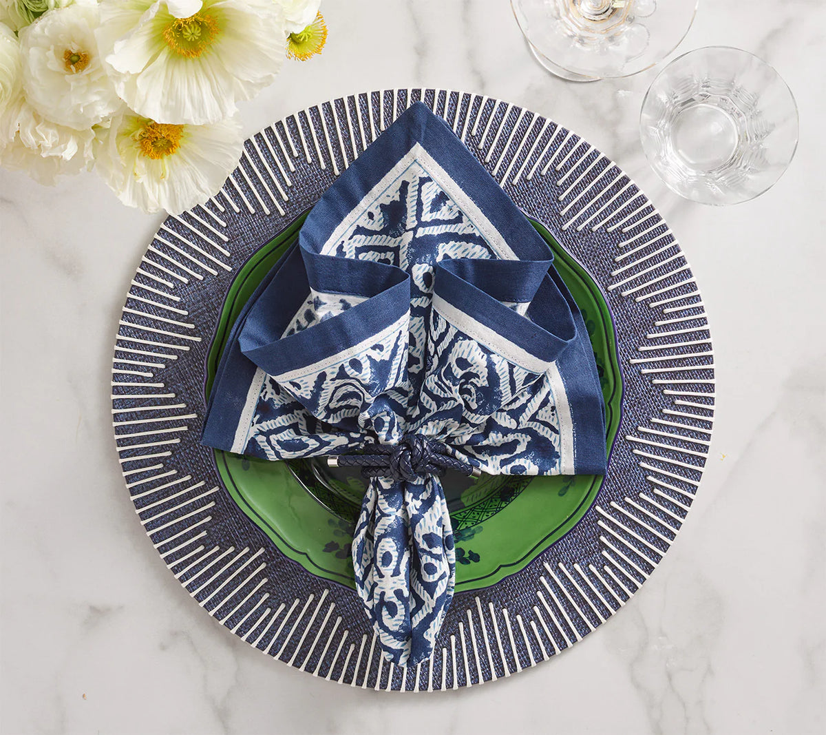 A blue and white napkin is folded on a green plate, set atop Kim Seybert’s Dream Weaver Placemat in navy & white. Nearby, two glasses and white flowers rest on the marble surface.