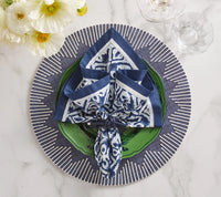 A blue and white napkin is folded on a green plate, set atop Kim Seybert’s Dream Weaver Placemat in navy & white. Nearby, two glasses and white flowers rest on the marble surface.