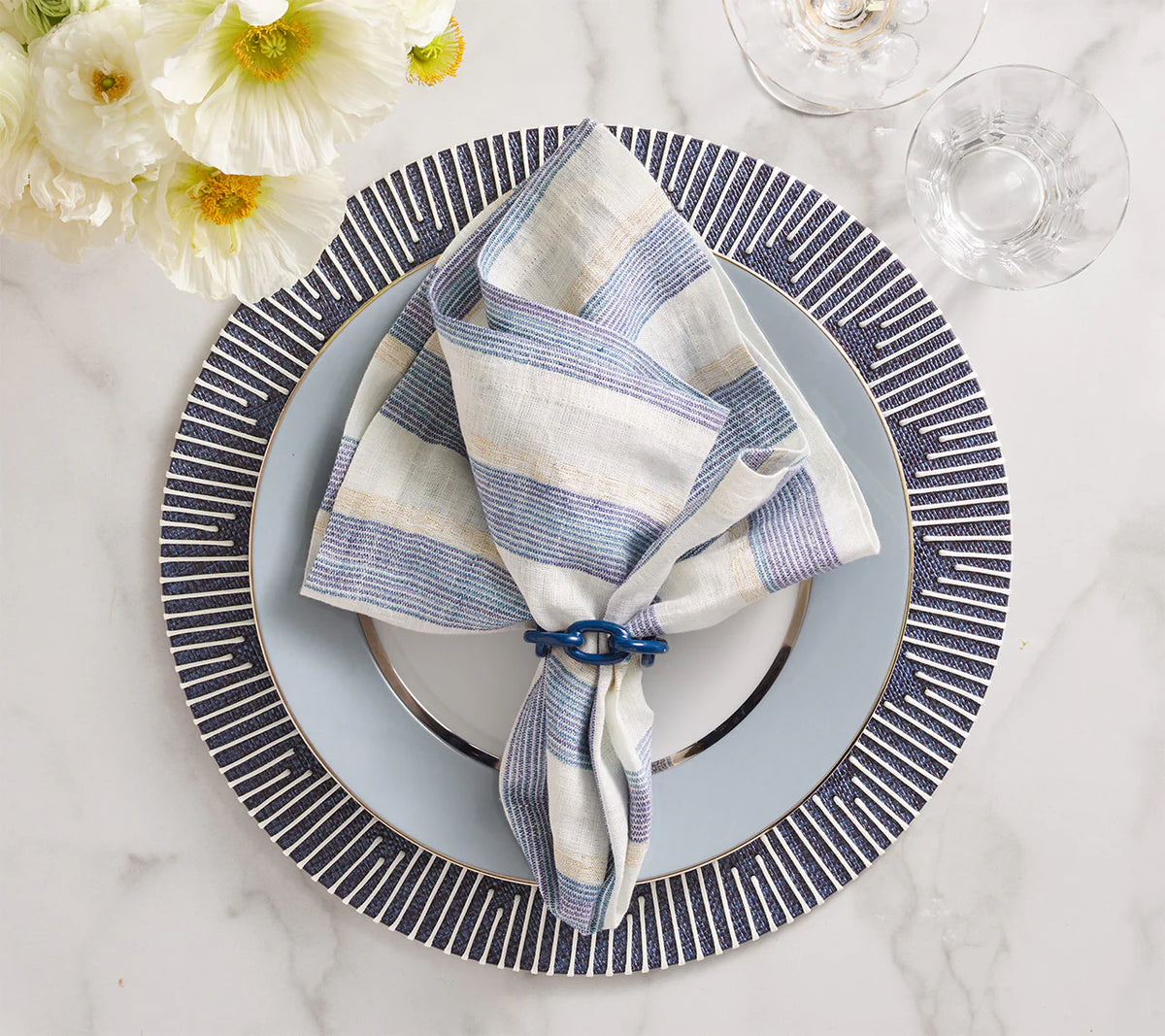 A striped linen napkin in blue and beige rests in a napkin ring atop a navy and white Dream Weaver Placemat by Kim Seybert, set on a patterned charger. Glassware and white flowers accent the marble tabletop.