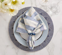 A striped linen napkin in blue and beige rests in a napkin ring atop a navy and white Dream Weaver Placemat by Kim Seybert, set on a patterned charger. Glassware and white flowers accent the marble tabletop.