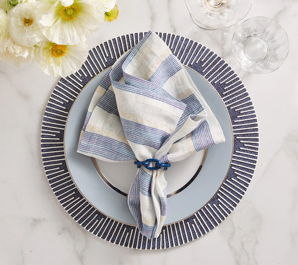 A striped linen napkin in blue and beige rests in a napkin ring atop a navy and white Dream Weaver Placemat by Kim Seybert, set on a patterned charger. Glassware and white flowers accent the marble tabletop.