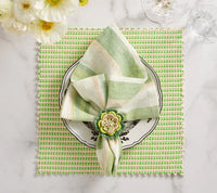A green and white striped napkin with a floral ring rests on a decorative plate atop the Kim Seybert Duet Placemat Set of 4 - Blue & Green. White flowers and glassware add elegance to these sophisticated table settings on marble.