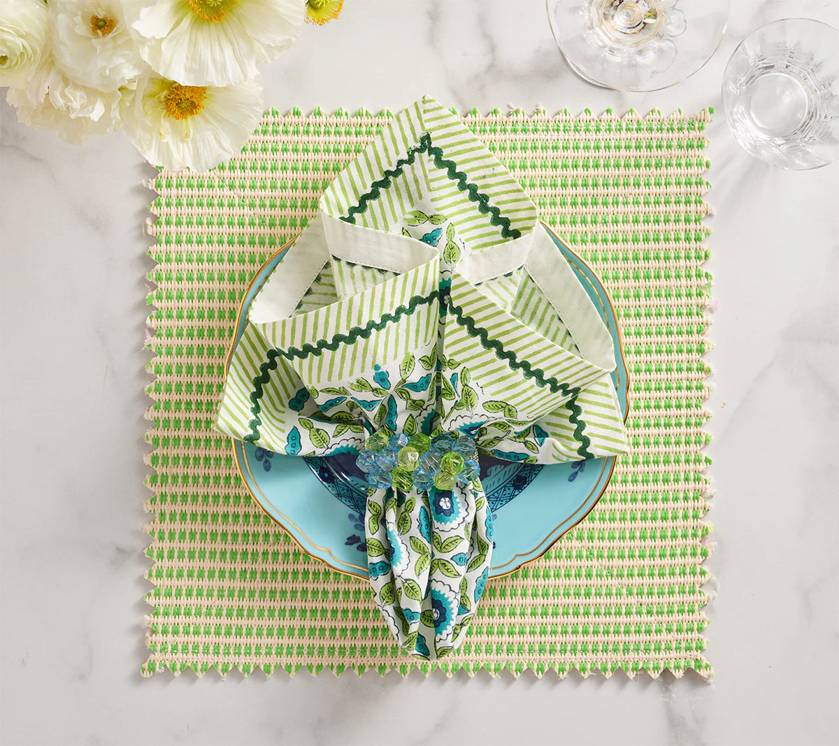 A tulip-shaped napkin on a blue floral plate rests atop the Kim Seybert Duet Placemat Set of 4 - Blue & Green, with white flowers and glassware enhancing the elegant marble table setting.