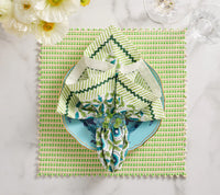 A tulip-shaped napkin on a blue floral plate rests atop the Kim Seybert Duet Placemat Set of 4 - Blue & Green, with white flowers and glassware enhancing the elegant marble table setting.