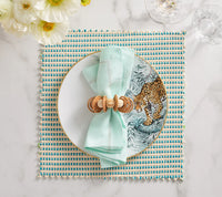 A table setting with a white plate and leopard design, a light blue napkin with decorative ring, Kim Seybert's Duet Placemat Set of 4 in Blue & Green for pattern and flair, plus white flowers on marble—perfect for elegant dining.