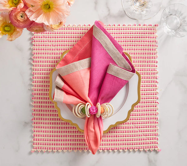 A stylish table features a pink and coral ombré fan-folded napkin with a ring on a white gold-edged plate, atop the Kim Seybert Duet Placemat Set of 4 in Blush & Pink. Flowers and glassware complete this elegant look.