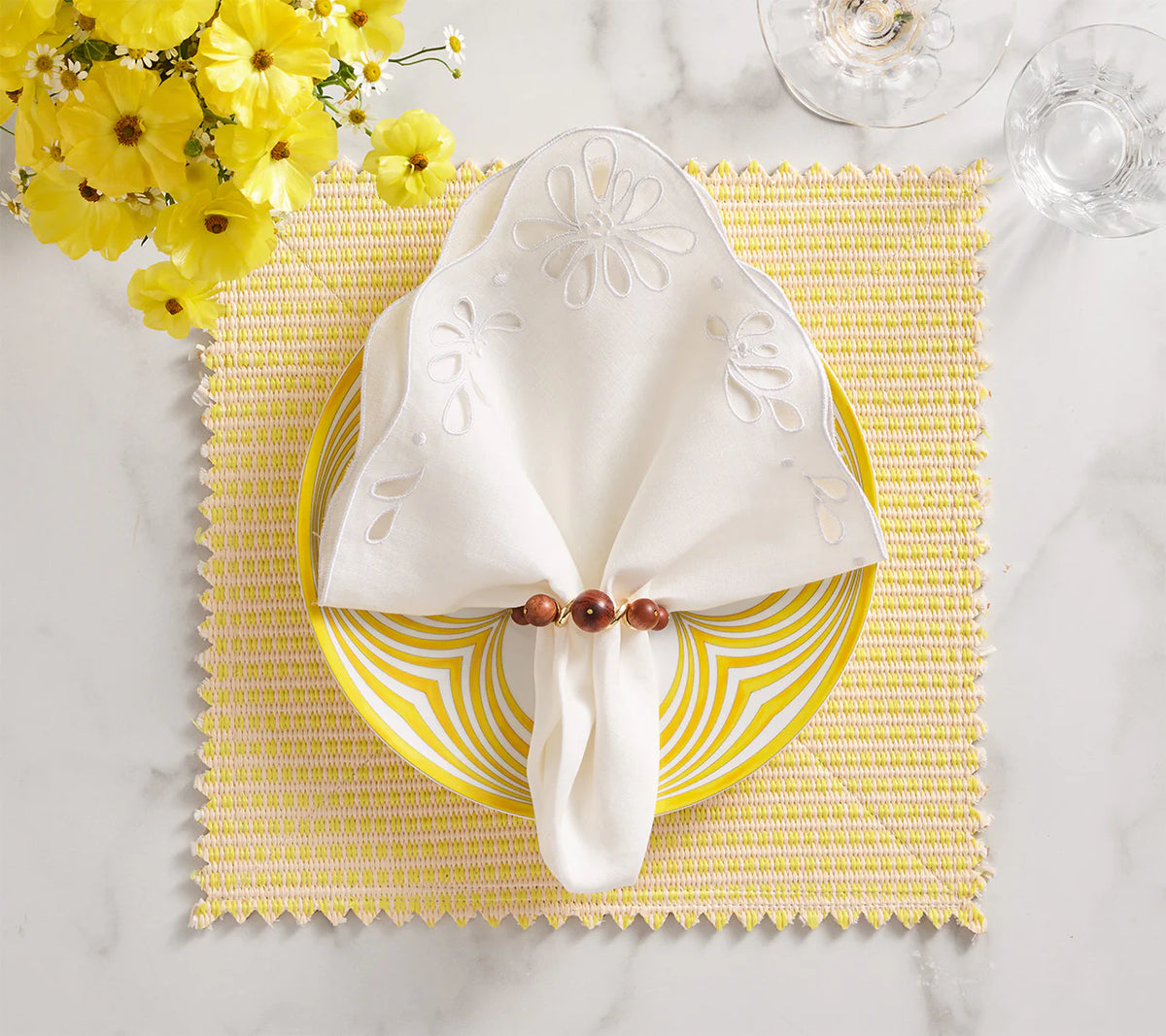 A table setting with a yellow and white striped plate, a white napkin held by Kim Seybert's Bois Napkin Ring Set of 4 in brown & gold, a yellow woven placemat, yellow flowers, and clear glassware on a white surface.