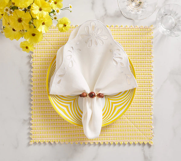 A table setting with a yellow and white striped plate, a white napkin held by Kim Seybert's Bois Napkin Ring Set of 4 in brown & gold, a yellow woven placemat, yellow flowers, and clear glassware on a white surface.