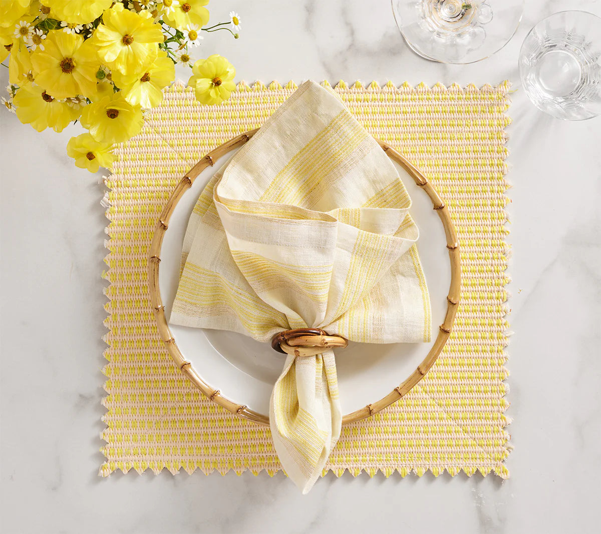 A coastal table setting with a white plate on a yellow and white placemat, Kim Seybert’s Broad Stripe Napkin Set of 4 - Yellow & White in wooden rings, wine glasses, and yellow flowers arranged on a white marble surface.