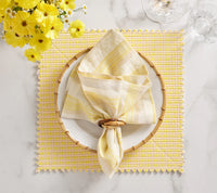 A coastal table setting with a white plate on a yellow and white placemat, Kim Seybert’s Broad Stripe Napkin Set of 4 - Yellow & White in wooden rings, wine glasses, and yellow flowers arranged on a white marble surface.