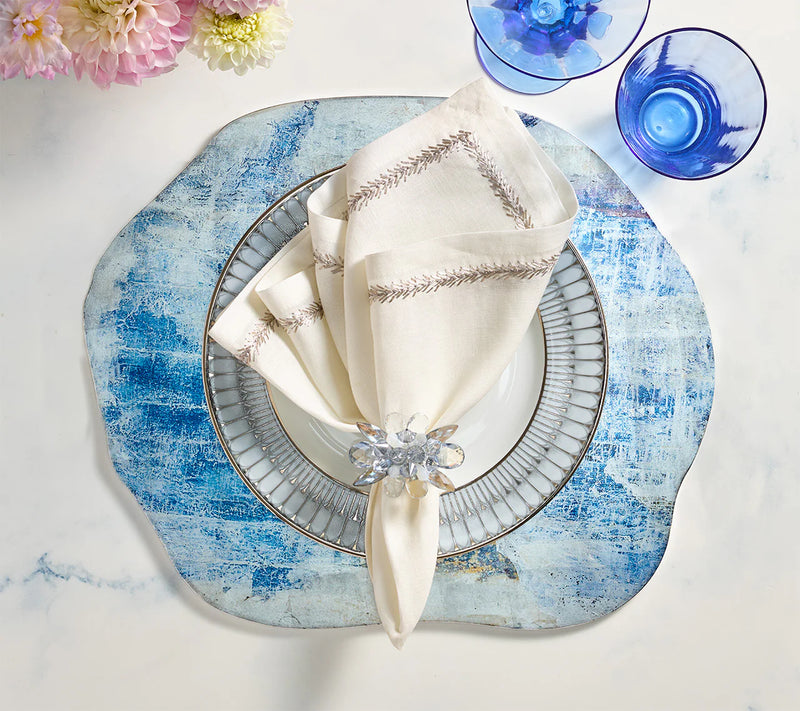 A white plate with a silver rim on a blue placemat, cream napkin with Kim Seybert Spectra Napkin Ring Set of 4 - Glacier Blue, blue glassware, and pink flowers creates elegant table décor on a white marble surface.