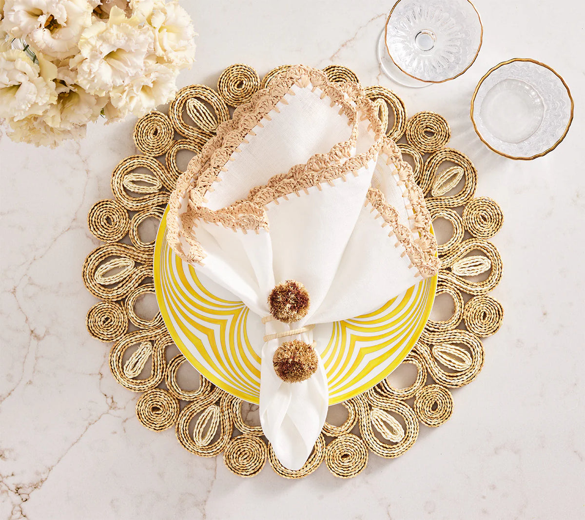 A decorative place setting features the Kim Seybert Marisol Placemat Set of 4 in Ivory & Natural, a yellow and white patterned plate, a beige-trimmed napkin with pom-poms, gold-rimmed glassware, and white flowers for natural table decor.