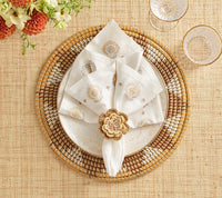 A decorative place setting featuring a woven placemat, a white plate with gold accents, a white floral napkin held by the Kim Seybert Pareo Napkin Ring Set of 4 in Natural & Brown, clear glasses, and flowers visible at the top left corner.