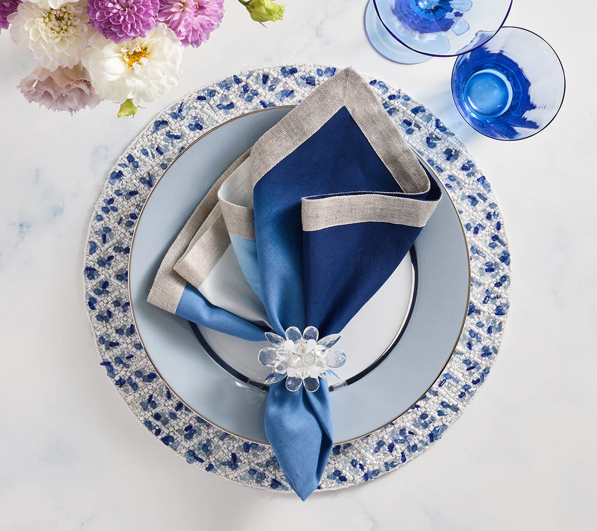 A gradient napkin held by the Kim Seybert Spectra Napkin Ring Set of 4 - Glacier Blue accents a blue and white place setting, with blue glassware and florals atop a decorative charger on a white marble table.