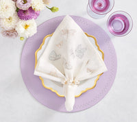 A white linen napkin with embroidered butterflies, held by Kim Seybert’s Pearl Drift Napkin Ring in pearl and gold, rests on a scalloped gold-rimmed plate atop a lavender placemat. White and purple flowers complete the elegant table setting.
