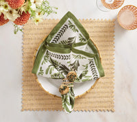 A white plate rests on a Kim Seybert Vista Reversible Placemat in Ivory & Beige, paired with a green-and-white leaf napkin, pom-pom napkin rings, and glassware, with flowers partially visible.