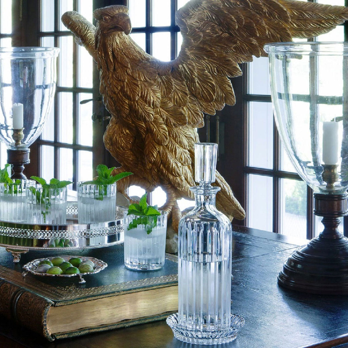 A decorative table displays a large gold eagle sculpture, tall glass vases, a Baccarat Mille Nuits wine coaster, mint cocktails on a silver tray, and a bowl of olives atop a large book before tall windowpanes.