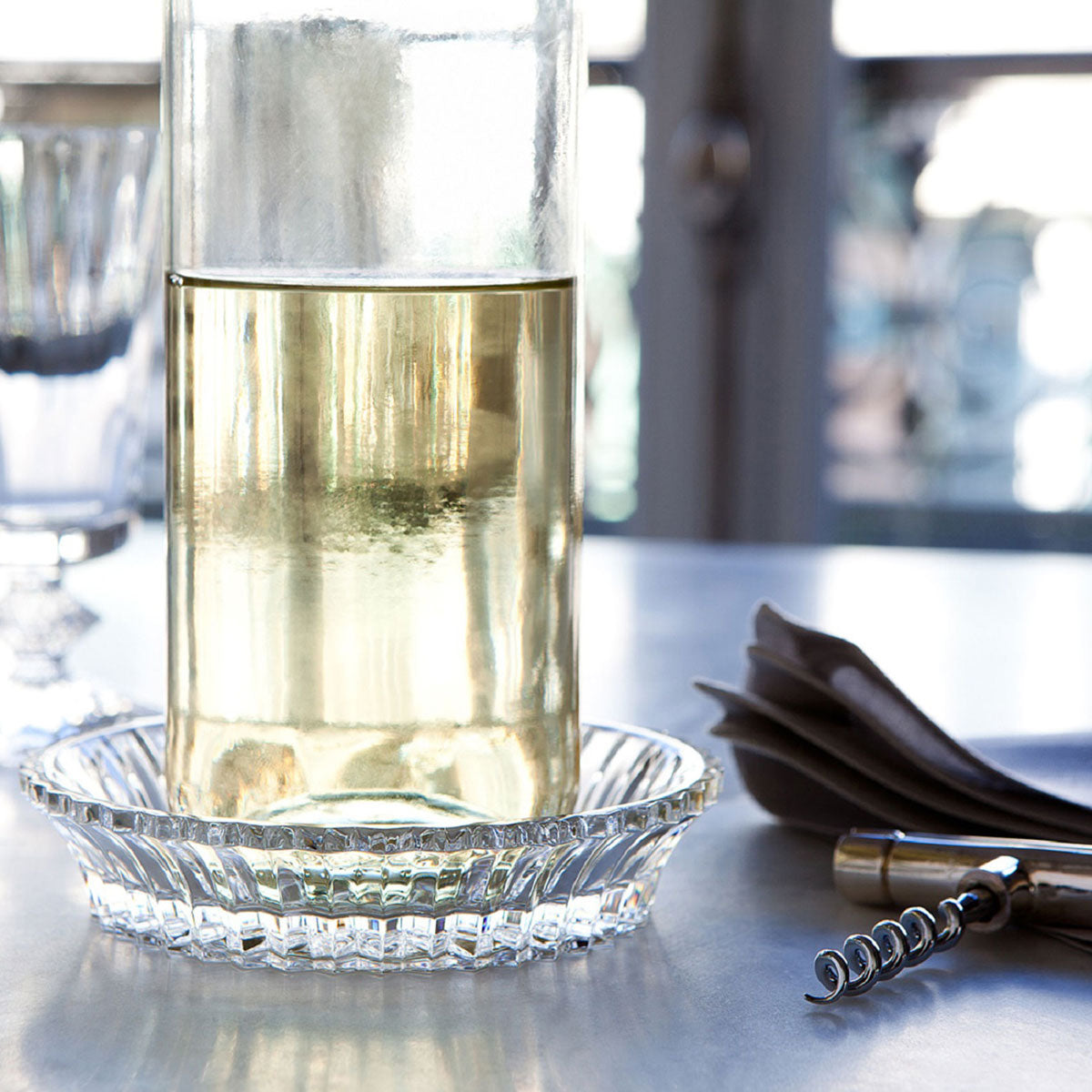 A glass bottle with yellow liquid rests on a Baccarat Mille Nuits Wine Coaster, accompanied by a corkscrew and folded napkins.