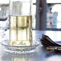 A glass bottle with yellow liquid rests on a Baccarat Mille Nuits Wine Coaster, accompanied by a corkscrew and folded napkins.