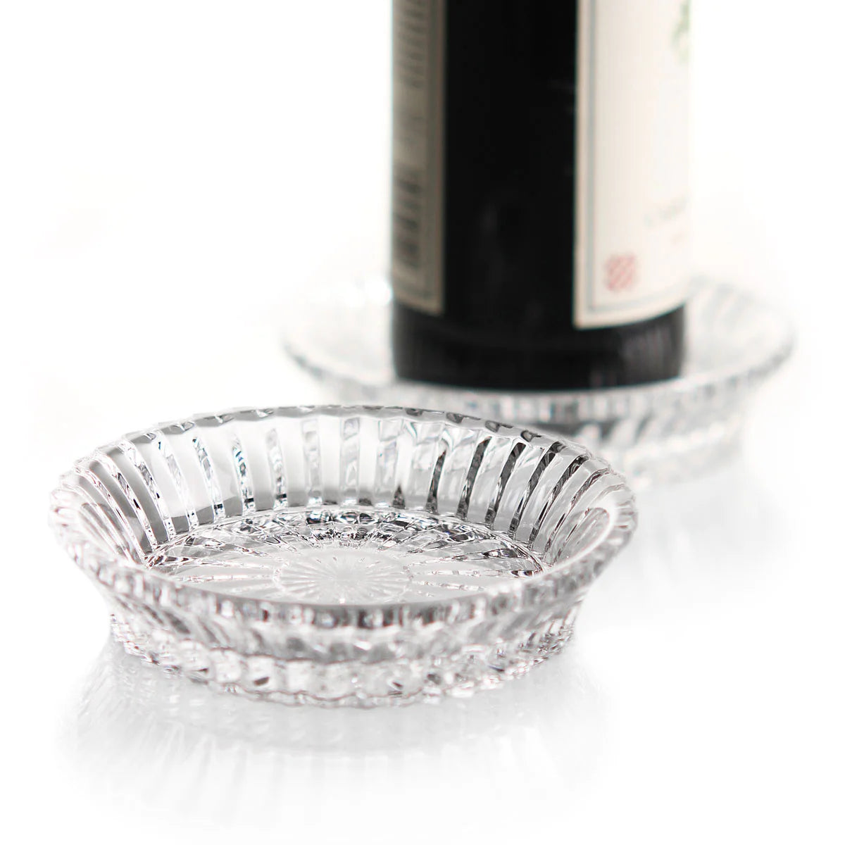 Two round Mille Nuits Wine Coasters by Baccarat, featuring a ridged design, rest on a white surface—one empty in the foreground and the other holding a dark crystal bottle in the background.