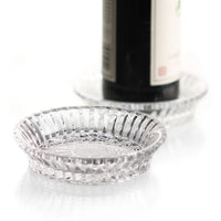 Two round Mille Nuits Wine Coasters by Baccarat, featuring a ridged design, rest on a white surface—one empty in the foreground and the other holding a dark crystal bottle in the background.
