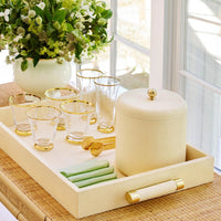 A cream-colored serving tray holds six gold-rimmed glasses, a Shagreen Ice Bucket by Aerin with gold tongs, green napkins, and a white vase of green and white flowers on a wicker table—perfect for an elegant home bar.