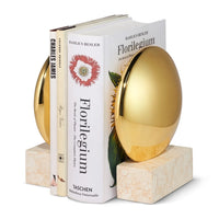The Aerin Constantin Bookend Set of 2 features marble bases and shiny gold egg-shaped accents in a mid-century style, holding three books—Florilegium, Charles James, and one other—upright against a plain white background.